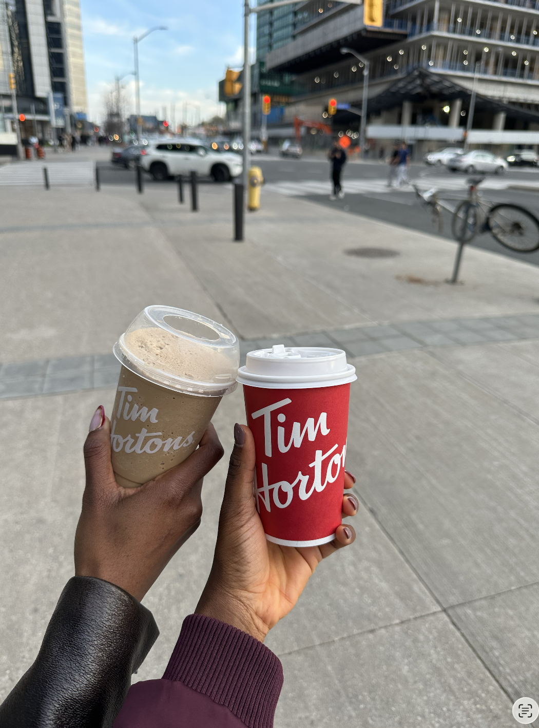 Two hands holding Tim Hortons drinks on a Toronto sidewalk with the city street in the background.